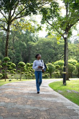 Genç bir kadın, zengin bir parkta kitap ve sırt çantası taşıyarak huzurlu bir gezintinin tadını çıkarıyor.