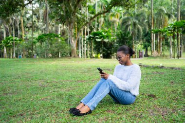 Parkta çimlerin üzerinde oturan gülümseyen siyah kadın.