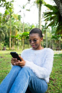 Tropikal bir parkta cep telefonu kullanan şık genç bir kadın, açık havada bağlantının tadını çıkarıyor.