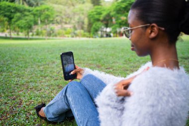 Parkta çimenlerde oturan genç bir kadın akıllı telefonuyla selfie çekiyor.