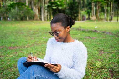 Genç siyahi kadın, parkta çimlerin üzerinde otururken defterine not alıyor.