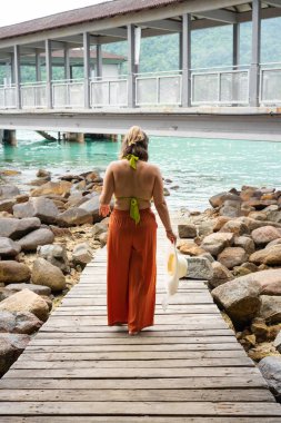 Genç bir kadın, tropik bir bungalova doğru ahşap bir iskelede yürüyor, rahatlatıcı bir tatilin tadını çıkarıyor.