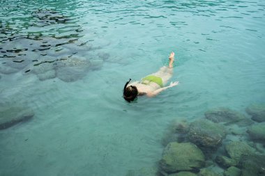 Turist kristal berrak turkuaz suda şnorkelle yüzüyor kayaların yanındaki deniz tabanını keşfediyor.