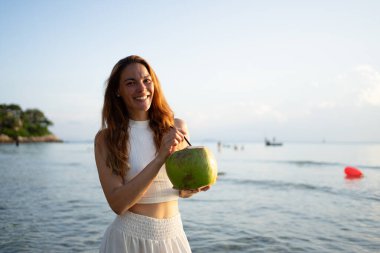 Gün batımında güzel bir sahilde ferahlatıcı bir hindistan cevizinin tadını çıkaran genç bir kadın.