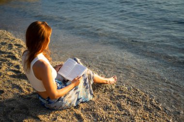 Gün batımında huzurlu bir plajda dinlenirken kitap okumaktan zevk alan bir kadın.
