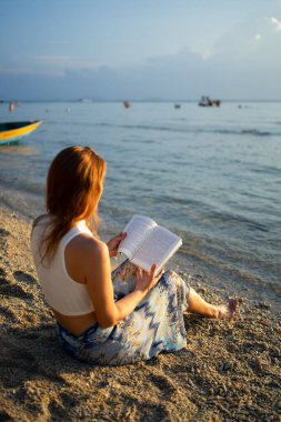 Gün batımında güzel bir plajda dinlenirken kitap okumaktan hoşlanan bir turist.