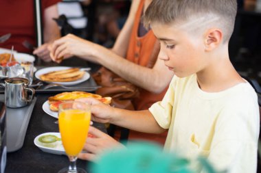 Genç bir çocuk kahvaltıda ailesiyle bir kafede tost yiyip portakal suyu içiyor.