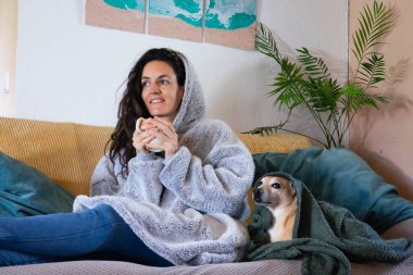 Kanepede evcil köpeğiyle dinlenirken, büyük boy kapüşonlu rahat bir içeceğin keyfini çıkaran bir kadın.