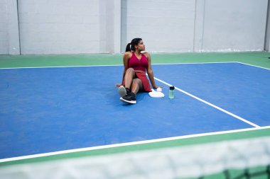 Hintli sporcu kadın. Pickleball antrenmanından sonra dinleniyor. Mavi iç sahada oturuyor.
