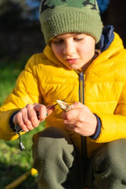 Sarı ceketli ve kamuflajlı bir çocuk. Şapka yontma çubuğu ve çakı.