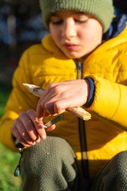 Küçük bir bıçakla bir tahta parçasını şekillendirmeye odaklanan genç bir çocuk, açık hava becerilerini öğreniyor.