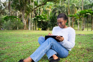 Odaklanmış kız öğrenci, parkta çimlerin üzerinde otururken defterine not alıyor, temiz havanın ve huzurlu çevrenin tadını çıkarıyor.