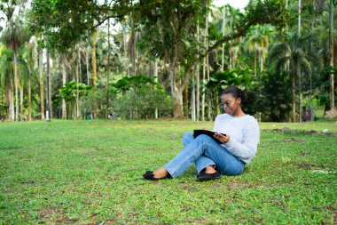 Üniversite öğrencisi açık havada ders çalışıyor. Not defterine not alıyor. Tropikal bir parkta çimlerin üzerinde oturuyor.