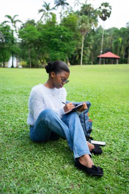 Genç bir kadın parkta çimlerin üzerinde oturmuş, temiz havanın ve yeşilliğin tadını çıkarırken deftere not alıyor.