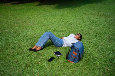 Üniversite öğrencisi okuduktan sonra çimenlerde dinleniyor, kampüs parkında tatilin tadını çıkarıyor.
