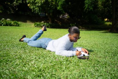 Üniversite öğrencisi parkta çimlerin üzerinde güneşli havanın ve temiz havanın tadını çıkarırken notlar alıyor.