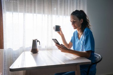 Mavi önlüklü doktor mutfaktaki akıllı telefonu kontrol ederken sabah kahvesinin tadını çıkarıyor.