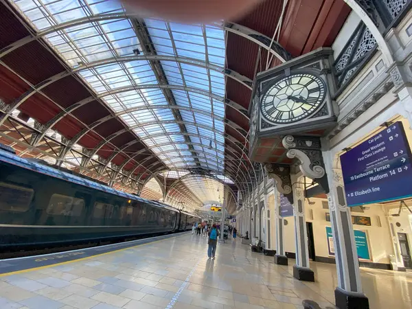 Londra 'nın Paddington İstasyonu' ndaki platformun geniş açılı, düşük ışık görüntüsü. Simgesel saat merkezi bir özelliktir, cam ve metal çatı yapısı ise öğleden sonra güneşinin sıcacık ışığıyla parlar. Görüntü hareketli atmosferi iletir 