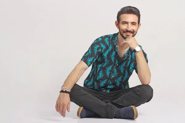 Genç İspanyol bir adam yerde oturuyor. Kendinden emin ve mutlu görünüyor. Gülümsüyor, kolları ve elleri çenesinde. Olumlu düşünmek.