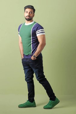 full length view of handsome young man in green shirt standing with arms crossed and looking at camera on grey background clipart