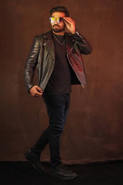 young man with leather jacket and sunglasses posing in studio clipart