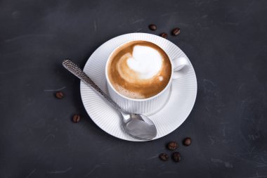 Üzerinde metal kaşık ve süt köpüğü olan siyah masada bir fincan kahve..