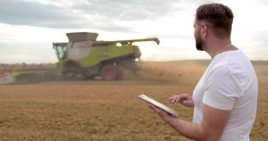 Tarım. Bir tarım uzmanı buğday tarlasında bir tabletle ayakta duruyor, arka planda bir kombine çalışıyor. Bir biçerdöver çiftçinin tarlasında buğday biçer..