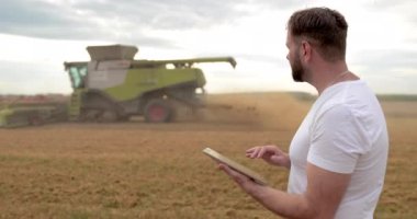 Tarım. Bir tarım uzmanı buğday tarlasında bir tabletle ayakta duruyor, arka planda bir kombine çalışıyor. Bir biçerdöver çiftçinin tarlasında buğday biçer..