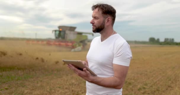 農業について アグロノミストは小麦畑にタブレットを置いており バックグラウンドにはコンバイン作品があります ハーベスターは農家の畑で小麦を収穫する — ストック動画