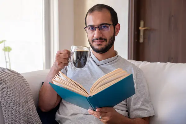 Bearded arab man drinking coffee while reading book and sitting on living room couch