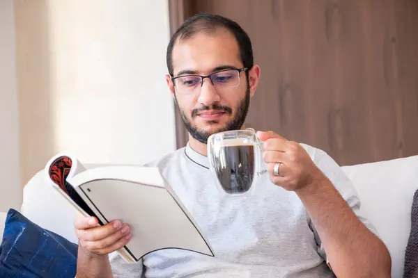 Bearded arab man drinking coffee while reading book and sitting on living room couch