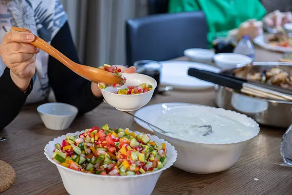 The wife fills a salad bowl with a large bowl of yogurt next to it to provide the guests with what they need at lunchtime at the large collective table for the large family.