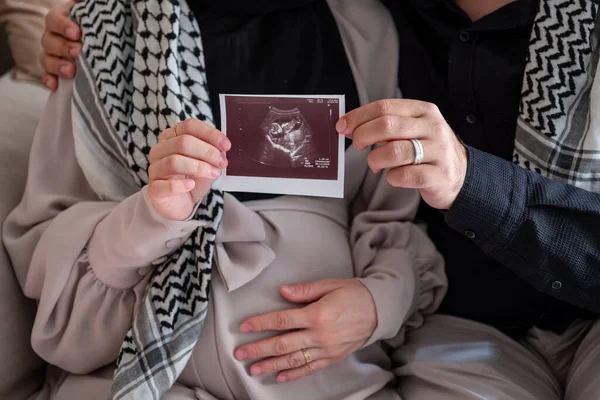 Husband and his wife holding sonography photo for there fetus while wearing palestinian keffiyeh in living room