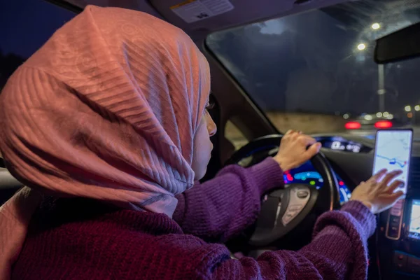 arabic muslim woman driving car at night wearing winter clothes ,hijab and seat belt driving in confidently and using maps through her mobile phone and GPS