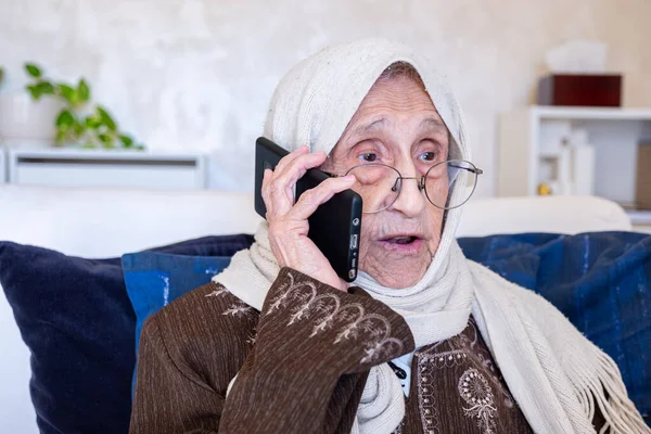 old woman wearing her glasses talking through her smartphone in modern living room, old woman making phone call