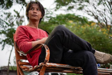 Kırmızı gömlekli bir Cava 'lı, bankta oturuyor, arka planda dikiliyordu. Düşük açılı bir bakış açısı vardı.. 