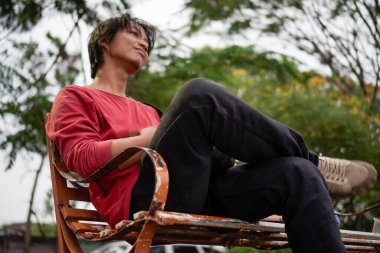 Kırmızı gömlekli bir Cava 'lı, bankta oturuyor, arka planda dikiliyordu. Düşük açılı bir bakış açısı vardı.. 