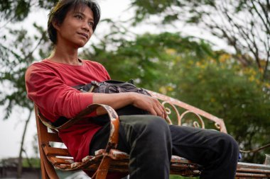 Kırmızı gömlekli bir Cava 'lı, bankta oturuyor, arka planda dikiliyordu. Düşük açılı bir bakış açısı vardı.. 