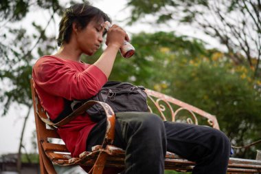 Kırmızı gömlekli bir Cava 'lı, bankta oturuyor, arka planda dikiliyordu. Düşük açılı bir bakış açısı vardı.. 