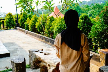 Arkadan bakıldığında, siyah başörtüsü takan bir kadın oturmuş doğal manzaraya bakıyor. 