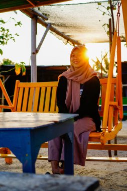 Ön taraftan başörtüsü takan bir kadın görünüyor. Dubangku 'da oturuyor. Arkasında güneş ışığı var.. 