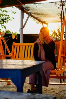 Ön taraftan başörtüsü takan bir kadın görünüyor. Dubangku 'da oturuyor. Arkasında güneş ışığı var.. 