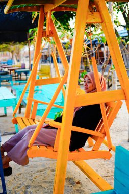 Ön taraftan başörtüsü takan bir kadın görünüyor. Dubangku 'da oturuyor. Arkasında güneş ışığı var.. 