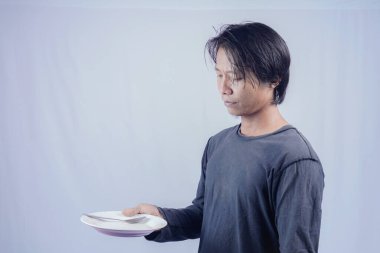 Yakışıklı Asyalı adam elinde boş bir tabak tutuyor, ne yiyeceği konusunda kafası karışık. Reklam için izole edilmiş beyaz çimen arka planı.