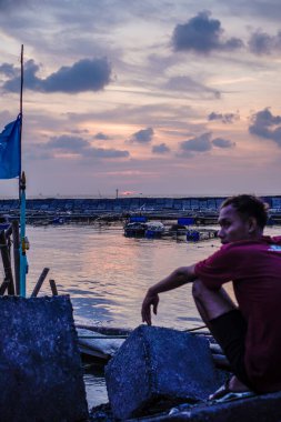 Jepara, Central Java, 7 Nisan 2024 - Cava 'lı adam denizin kenarında oturuyor bir dalgakıranın ve alacakaranlıkta gökyüzünün önünde balık tutuyor.