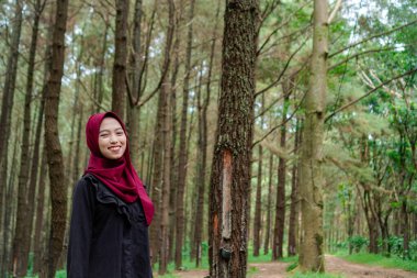 Başörtüsü takan Cava 'lı kadın ormanın ortasında kameraya bakıyor. Arka planda boş fotokopi alanı olan ağaçlar var.