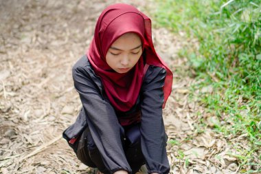 Asyalı kadın tesettürlü kayalık bir yolda kasvetli bir ifadeyle çömeliyor.