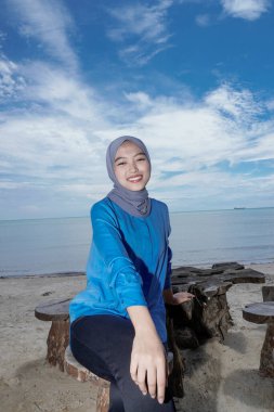 Başörtüsü takan bir kadın sahilde oturuyor. Parlak bir gökyüzüne karşı. Reklam için boş bir alanı var..