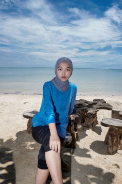 Başörtüsü takan bir kadın sahilde oturuyor. Parlak bir gökyüzüne karşı. Reklam için boş bir alanı var..