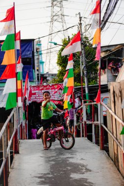 Ancol, Kuzey Jakarta, 10 Ağustos 2024, çocuklar nehir köprüsünde renkli bayrak direkleriyle bisiklet oynuyorlar..